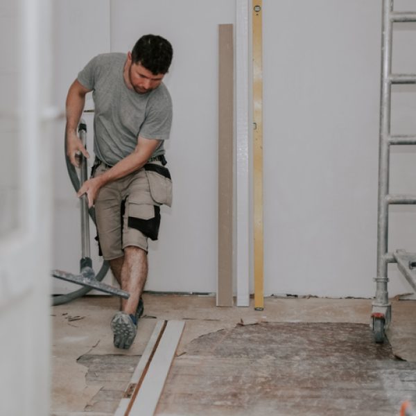 Young caucasian brunette man in work clothes on the left is vacuuming the floor with a construction vacuum cleaner in a room in an old house being renovated, side view close-up with selective focus.Construction work concept.