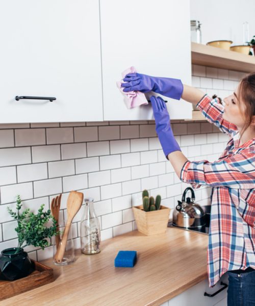 Woman in gloves cleaning cabinet with rag at home kitchen